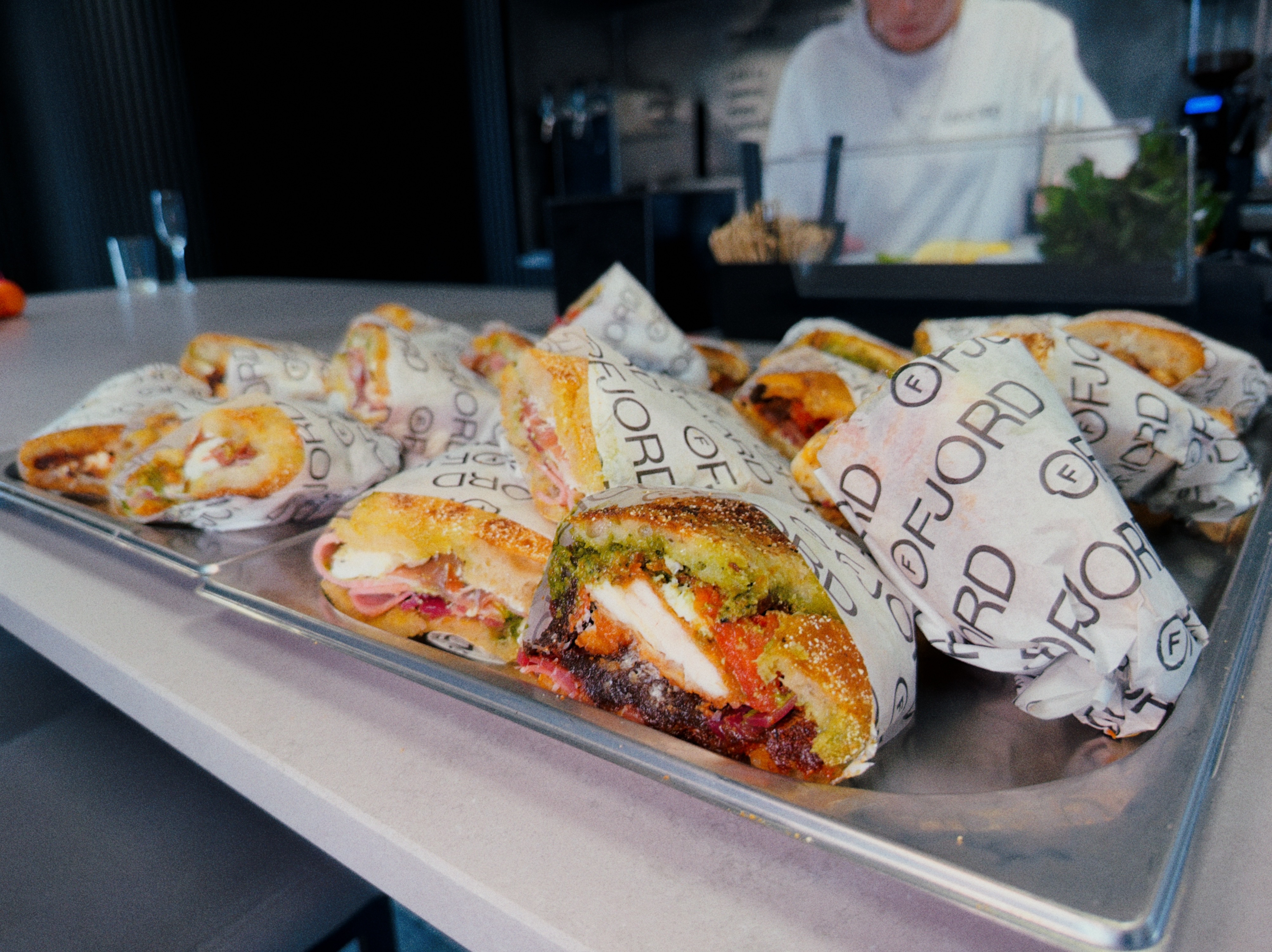 A full tray of Ruger Fjord focaccia sandwiches with bold fillings, Kieran in chef whites visible behind the counter.